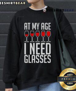 Woman wearing 'Nice At My Age I Need A Glass of Wine' sweatshirt, enjoying a cozy evening with a glass of wine.