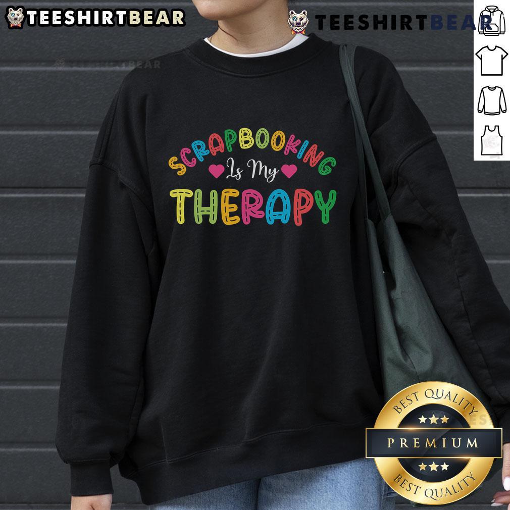 Cozy 'Pretty Scrapbooking Is My Therapy' sweatshirt displayed on a wooden table with scrapbook supplies.