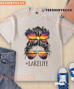 Women in a messy bun wearing 'Lake Life' washed t-shirts, enjoying a sunny day by the lake with family and friends.
